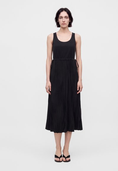 Woman standing wearing a sleeveless black top and pleated black midi skirt with black open-toe sandals against a plain light background.