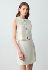 Geknipt wit tweed vest met twee voorzakken en gouden knopen, gecombineerd met een bijpassende rok. Gelaagde stof met een zachte afwerking.