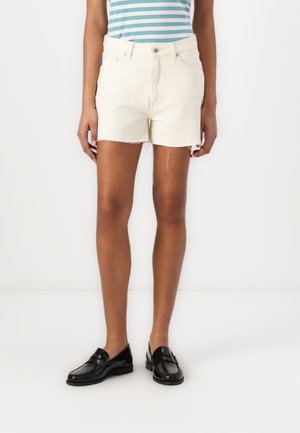 Personne portant un short en denim couleur crème avec des ourlets effilochés, un haut rayé bleu et blanc, et des mocassins noirs, debout devant un fond blanc.