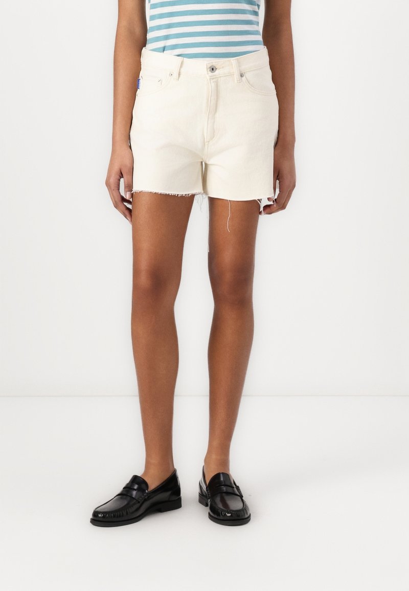 Personne portant un short en denim couleur crème avec des ourlets effilochés, un haut rayé bleu et blanc, et des mocassins noirs, debout devant un fond blanc.