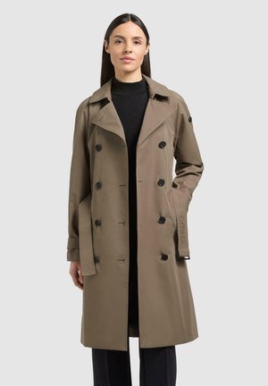 Kvinne iført en brun knelang dobbelspent trenchcoat over en svart høyhalset genser, stående mot en lys grå bakgrunn.