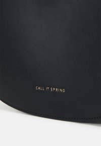 Gros plan d'un sac en cuir noir avec le logo doré « CALL IT SPRING » embossé près du bord inférieur, posé sur une surface blanche.