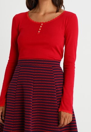 Kobieta ubrana w czerwoną bluzkę z długim rękawem z guzikami i falbaniastymi brzegami, w połączeniu z wysokim stanem spódnicy w granatowo-czerwone paski.