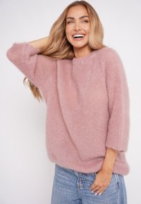 Uśmiechająca się kobieta z długimi blond włosami, ubrana w puszysty różowy sweter i jasnoniebieskie dżinsy, pozująca z jedną ręką za głową.