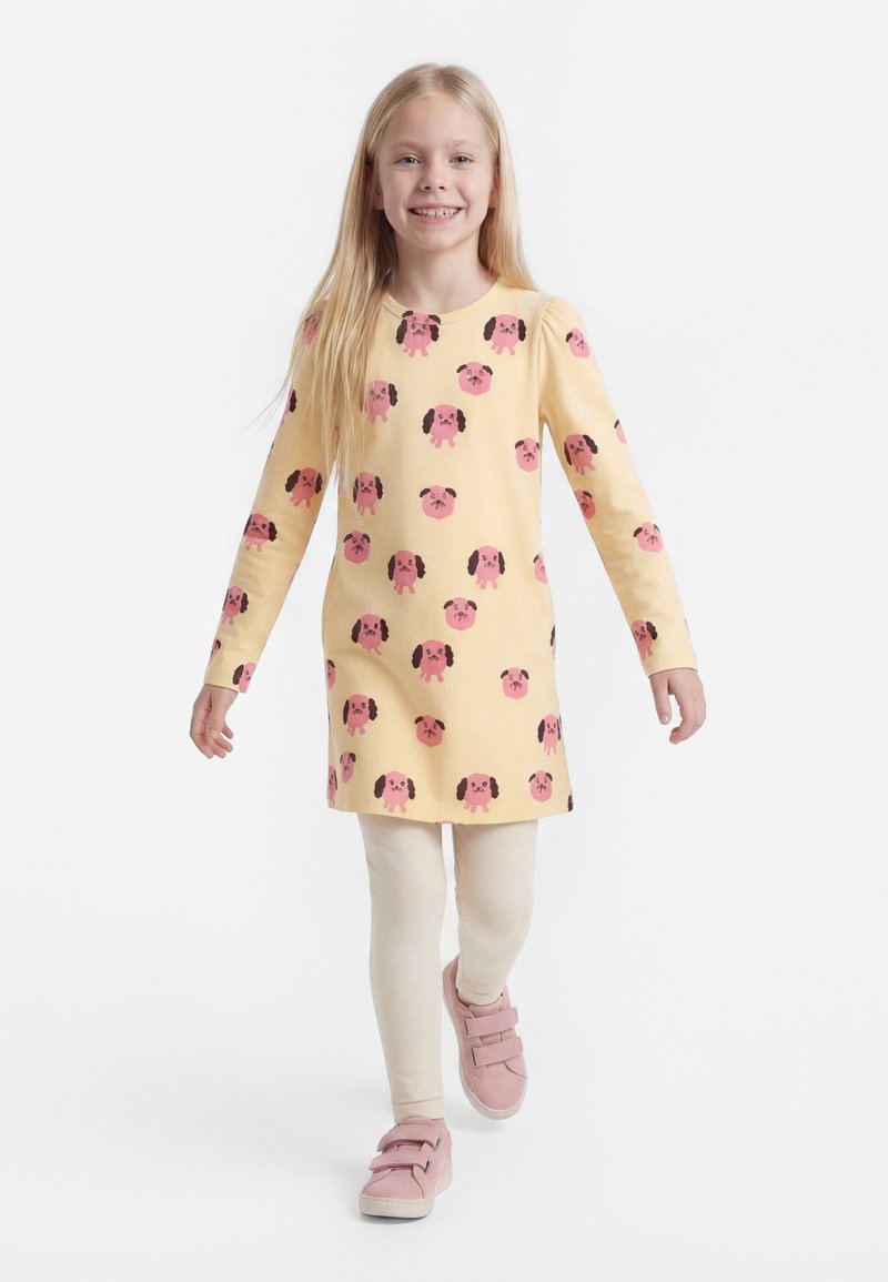 Lächendes Mädchen, das ein gelbes Kleid mit pinkfarbenen Hundegesichtern, cremefarbene Leggings und pinke Klettverschluss-Sneaker trägt und auf weißem Hintergrund läuft.