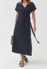 Granatowa sukienka kopertowa z krótkimi rękawami, dekoltem w serek, wiązaniem z boku i spódnicą sięgającą do ziemi; zestawiona z brązowymi sandałami z odkrytymi palcami.