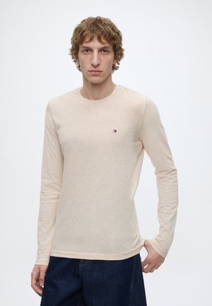 Giovane uomo con capelli ricci che indossa una camicia beige a maniche lunghe con un piccolo logo sul petto e jeans blu scuro su uno sfondo bianco semplice.
