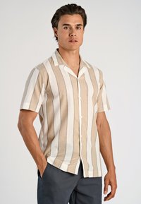 Man in een beige en witte verticaal gestreepte korte mouw button-up shirt met donkere broek, staand met één hand in de zak tegen een effen achtergrond.