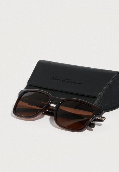 Occhiali da sole con montatura quadrata in tartaruga scuro e lenti marroni. Include una custodia nera texturizzata con logo in rilievo.
