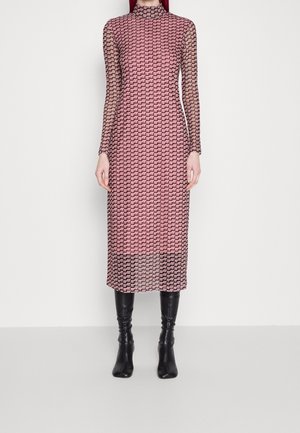 Frau trägt ein langärmeliges Midikleid mit einem geometrischen Muster in Pink und Schwarz, kombiniert mit schwarzen kniehohen Stiefeln, und steht vor einem weißen Hintergrund.