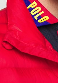 Röd puffjacka med en synlig blå krage som har gul "POLO"-text. Tyget har en slät, glänsande yta och tjock isolering.