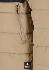 Beige pufferjacka med en svart övre del, med en sidoficka med dragkedja och en svart logotypmärke med ett bergsmotiv. Slät textur.
