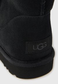 Svart ankelstövel i mocka med mjuk textur, utrustad med en UGG-logopatch och en flexibel svart gummisula. Sömnadsdetaljer längs kanterna.