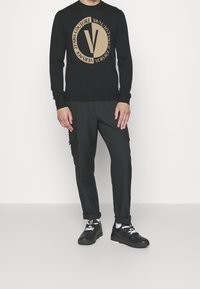 Pull noir avec cercle beige contrastant et logo "V", poignets côtelés, associé à un pantalon cargo noir et des baskets noires.