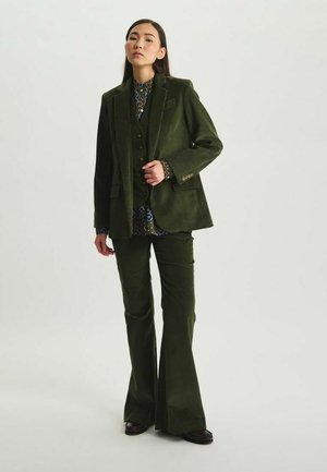 Costume en velours côtelé vert foncé avec une veste ajustée, un pantalon évasé et un chemisier à motifs. Les détails notables incluent des boutons et un tissu texturé.