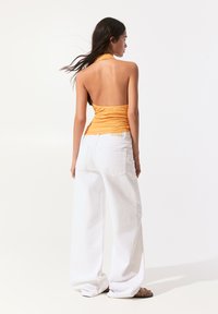 Oranžový halter top se shromážděnou texturou, spárovaný s volnými bílými džíny. Model zobrazen zezadu, minimalistické pozadí.
