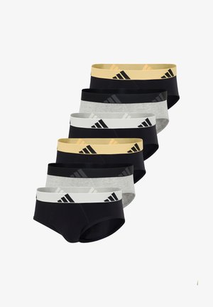 Athletischer Mann steht barfuß, trägt schwarze Slips mit gelbem Taillenbund und Adidas-Logo, schaut nach links mit einer Hand auf der Hüfte.