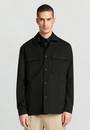 Giacca nera con bottoni e colletto, due tasche sul petto e una texture liscia. Abbinata a pantaloni beige.