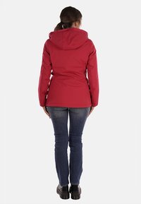 Giacca impermeabile rossa con cappuccio, design aderente, texture liscia e dettagli delle cuciture; abbinata a jeans scuri e stivali neri.