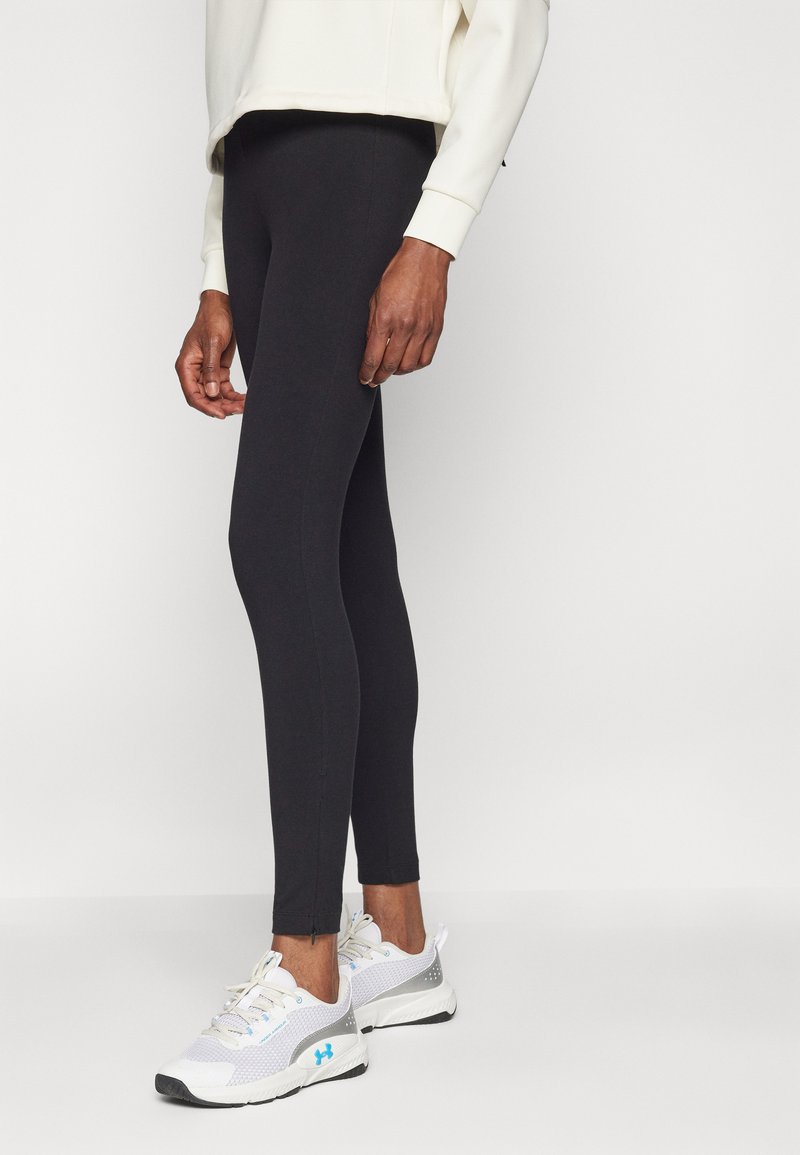 Leggings neri con un design aderente, realizzati in un materiale liscio e elasticizzato. Abbinati a scarpe da ginnastica bianche e grigie con accenti blu.