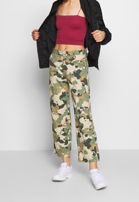 Kamouflage vida byxor i beige, grönt och brunt; ihop med en åtsittande marsalafärgad crop top och en svart bomberjacka; vita sneakers.