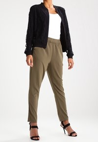 Giacca bomber in pizzo nero sopra una maglietta bianca, abbinata a pantaloni a sigaretta verde oliva e sandali neri con tacco alto.