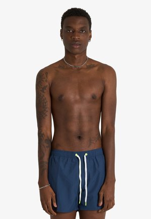 Giovane uomo con tatuaggi che indossa una collana a catena d'argento e pantaloncini da bagno blu con coulisse bianche e al neon, in piedi su sfondo bianco.
