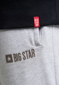 Šedé tepláky s hladkou textúrou, s výrazným logom "BIG STAR" v hnedej farbe a červenou značkovou etiketu na čiernom páse.
