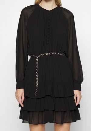 Zwarte jurk met lange mouwen en geplooide lagen, met stoffen knopen aan de voorkant, doorzichtige mouwen en een gouden kettingriem in de taille.