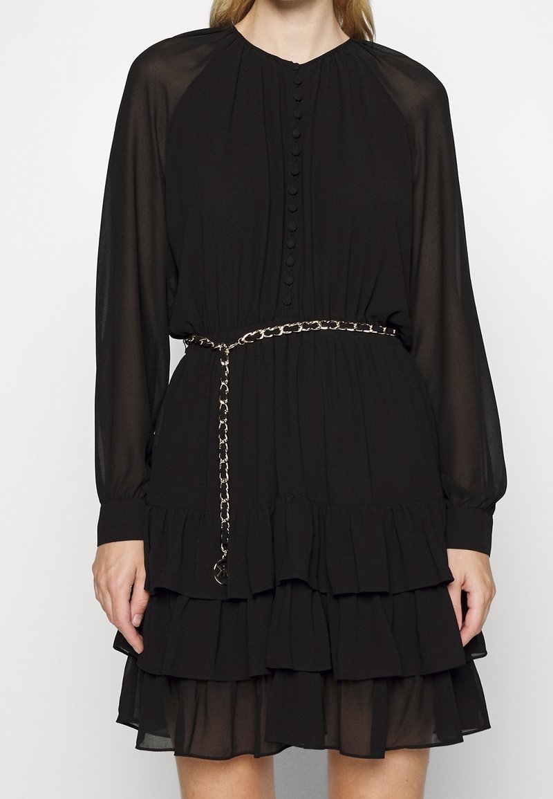Robe noire à manches longues avec volants en couches, boutons recouverts de tissu à l'avant, manches transparentes, et ceinture chaîne dorée à la taille.