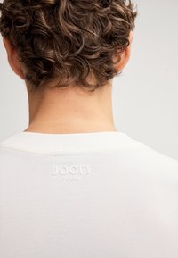 Weißes Sweatshirt mit geripptem Kragen. Auf der Rückseite befindet sich ein erhabenes Logo "JOOP! JEANS", das eine glatte Baumwollstruktur zeigt.