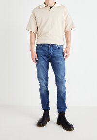 Beige kortärmad polo skjorta med vit krage, i kombination med blå denimjeans och svarta ankelskor med en texturerad sula.