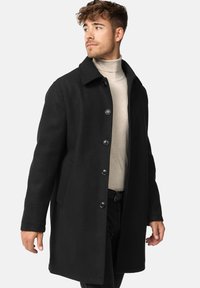 Sort uldfrakke med krave, lange ærmer og knapper foran. Har sidelommer og en lige pasform, båret over en beige turtleneck.