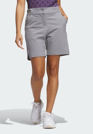 Kvinde iført lysegrå shorts, hvide sneakers og en lilla mønstret top, gående på en ensfarvet lys baggrund.