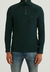 Tmavě zelený pletený svetr s vysokým límcem, předním zipem, žebrovanou strukturou a kontrastními švy na ramenou. Nosený s modrými džíny.
