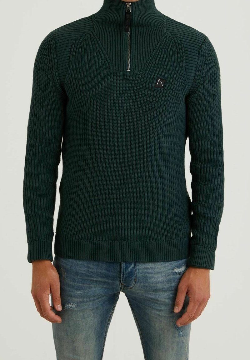 Tmavě zelený pletený svetr s vysokým límcem, předním zipem, žebrovanou strukturou a kontrastními švy na ramenou. Nosený s modrými džíny.