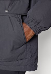 Dark gray puffer jacket with a ribbed cuff, featuring a side pocket and a brown leather glove partially visible within the pocket.