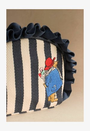 Triibuline kangast kotike tumesinise volangiga äärega ja tikitud karuga, kes kannab punast mütsi ja sinist mantlit ning kannab pintslit.