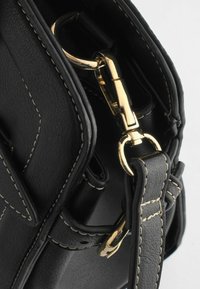 Bandoulière de sac en cuir noir avec des surpiqûres blanches attachée à un fermoir métallique doré et des anneaux sur un sac en cuir noir assorti.