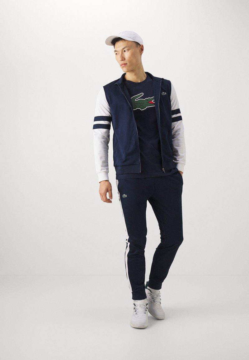 Ensemble de survêtement navy avec des accents blancs, comprenant un hoodie zippé et un pantalon assorti. Logo en caoutchouc sur le t-shirt, associé à des baskets blanches.