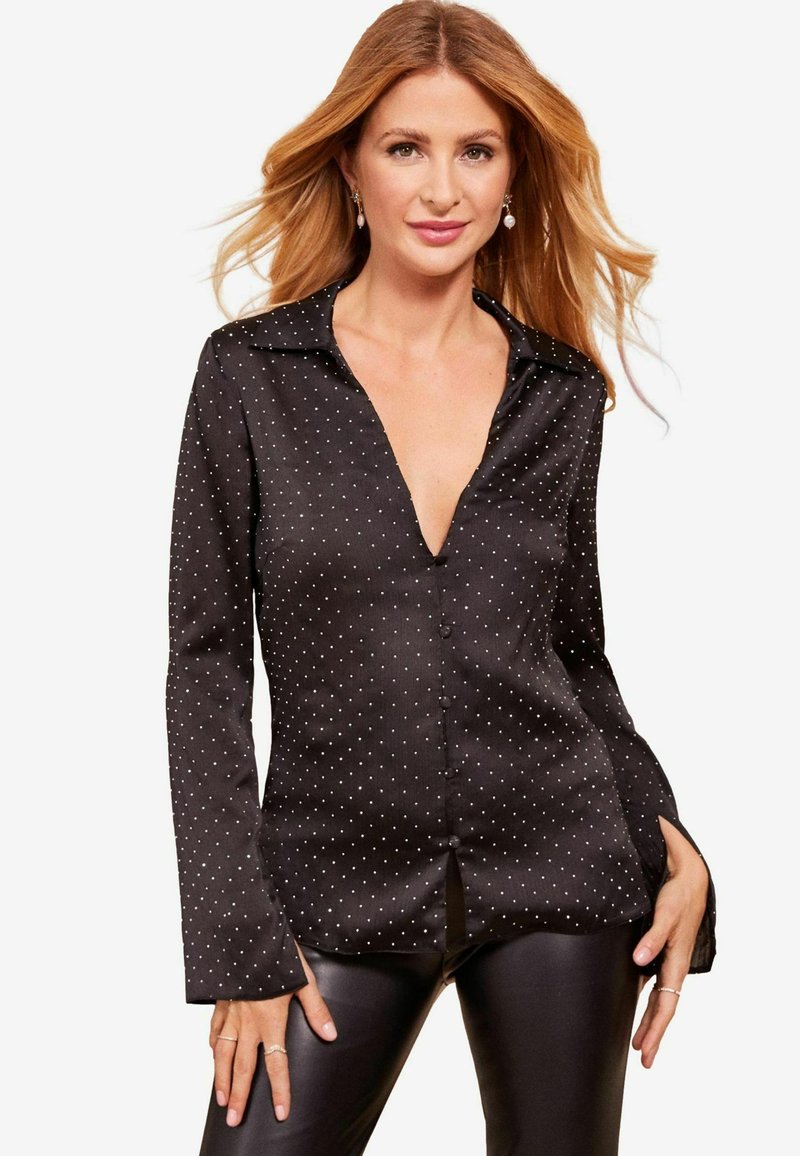 Blusa di raso nera con scollatura a V profonda, maniche lunghe e pois bianchi. Presenta una chiusura con bottoni sul davanti e polsini leggermente svasati.