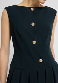 Robe sans manches vert foncé avec un corsage ajusté, une jupe évasée et des boutons dorés ornés sur le devant. Texture de tissu lisse.