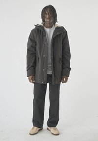 Mann mit schulterlangen Dreads trägt eine schwarze Kapuzenjacke über einem grauen Shirt, schwarze Hose und beigen Sneakers und steht vor einem schlichten Hintergrund.