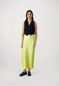 Femme debout portant un gilet sans manches noir sur une robe maxi en satin vert lime avec des mocassins noirs, fond clair uni.