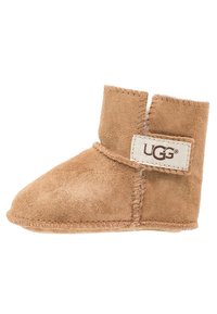Botín de ante marrón con una textura suave, punta redondeada y una correa de velcro. Presenta una pequeña etiqueta UGG en el lateral. Altura aproximada: tobillo.