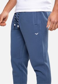 Modré bavlněné tepláky s elastickým páskem, bílým stahovacím šňůrkem, bočními kapsami a malým vyšívaným logem na levém stehně.