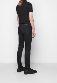 Jeans ajustados negros con una textura suave, un bolsillo trasero y una etiqueta con el logo. Combinados con una camiseta tipo polo negra y zapatillas negras.