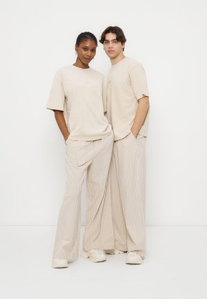 Δύο άτομα στέκονται δίπλα-δίπλα φορώντας μπεζ φαρδιές μπλούζες και κρεμ παντελόνια με λεπτές κάθετες ρίγες, πάνω σε απλό λευκό φόντο.
