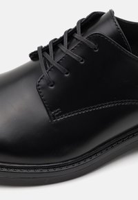 Chaussure habillée en cuir noir avec une finition lisse, un bout rond, un design à lacets et des détails de couture subtils le long du bord.