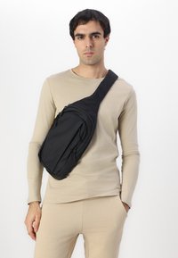 Svart stoffekse med diagonal bæring, som har flere glidelåslommer og en polstret stropp for komfort. Jevn tekstur.